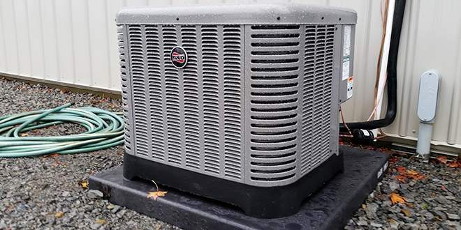 HVAC unit on a black base outside a metal building with green garden hose nearby.