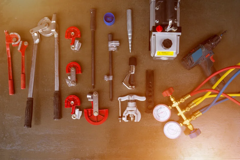 HVAC tools and equipment laid out on a dark surface, including a drill, pipe cutters, and colorful pressure gauges.