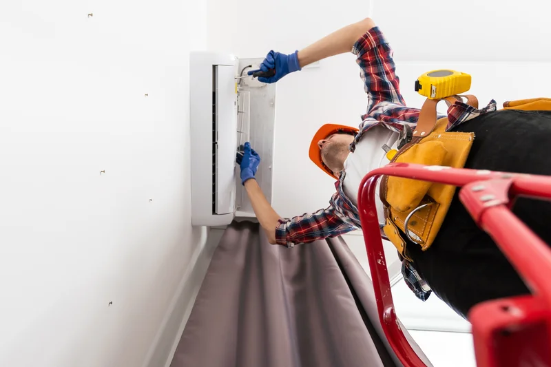 Technician in a hard hat and tool belt fixing an air conditioning unit on a ladder.