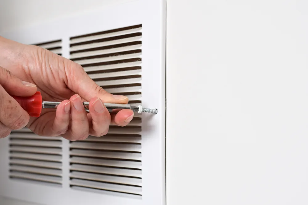 Hand using a red-handled screwdriver to adjust a screw on a white air vent.