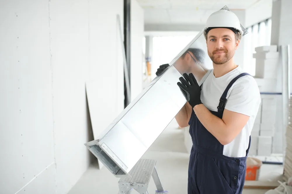 Worker in overalls and hard hat holding a large metal duct piece and smiling in a bright, unfinished room.