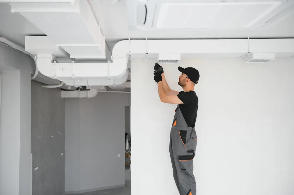 Technician in gray overalls installing an HVAC system on a white ceiling.