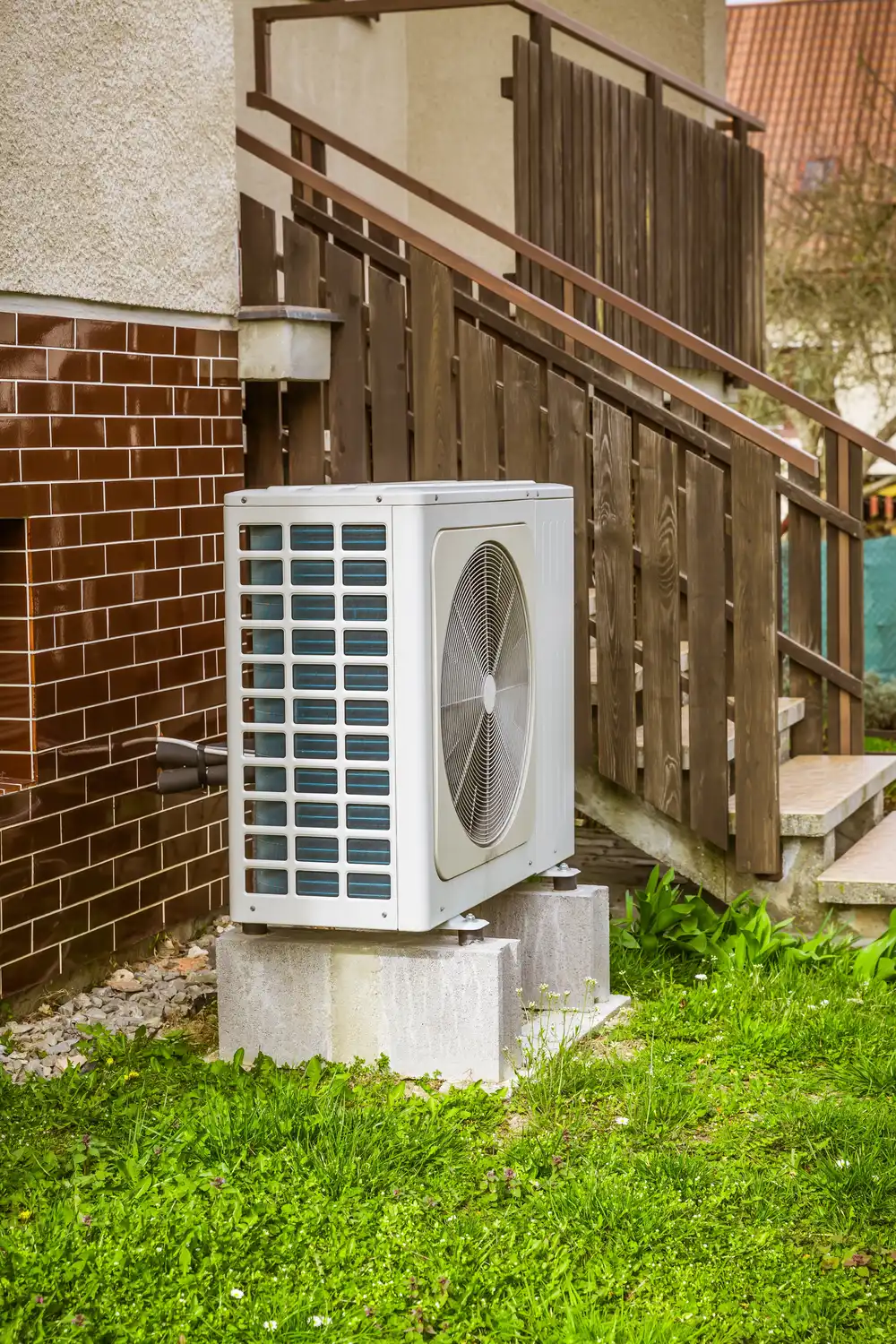 Outdoor HVAC unit on concrete blocks beside brick and wooden steps, surrounded by green grass.