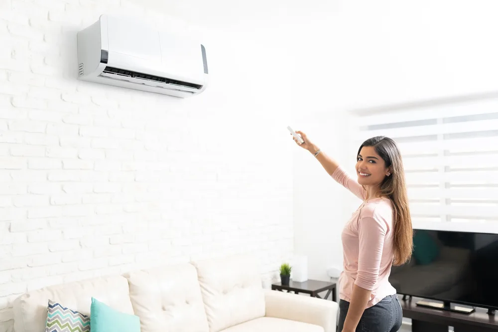 Smiling woman adjusting a wall-mounted air conditioner with a remote in a bright living room.