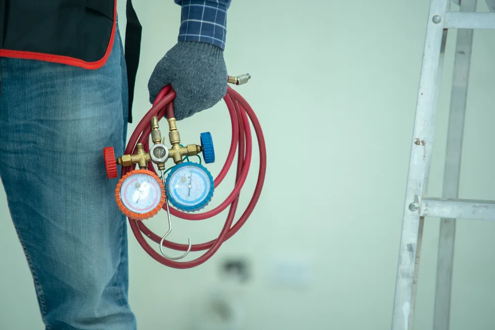 Technician in jeans holding HVAC gauges next to a ladder.