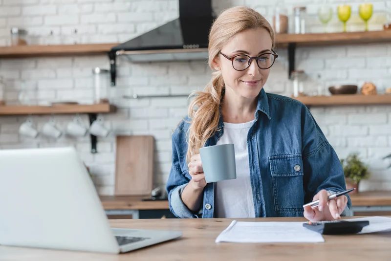 Woman in denim jacket holding a mug and working at a kitchen table with a laptop and paperwork.